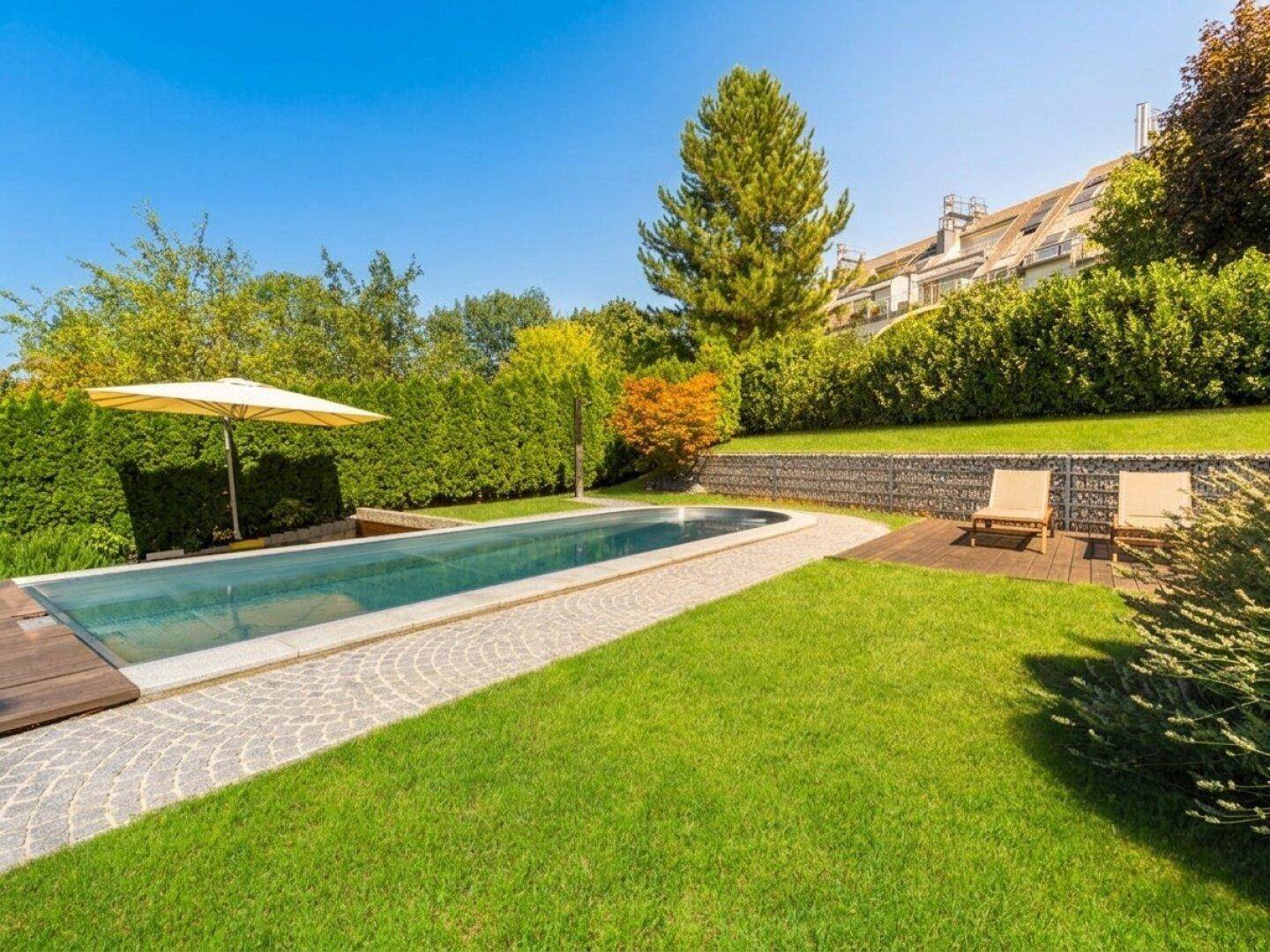 Sonnige Gartenanlage mit Pool, Liegewiese und Gabionenwand im Hintergrund.