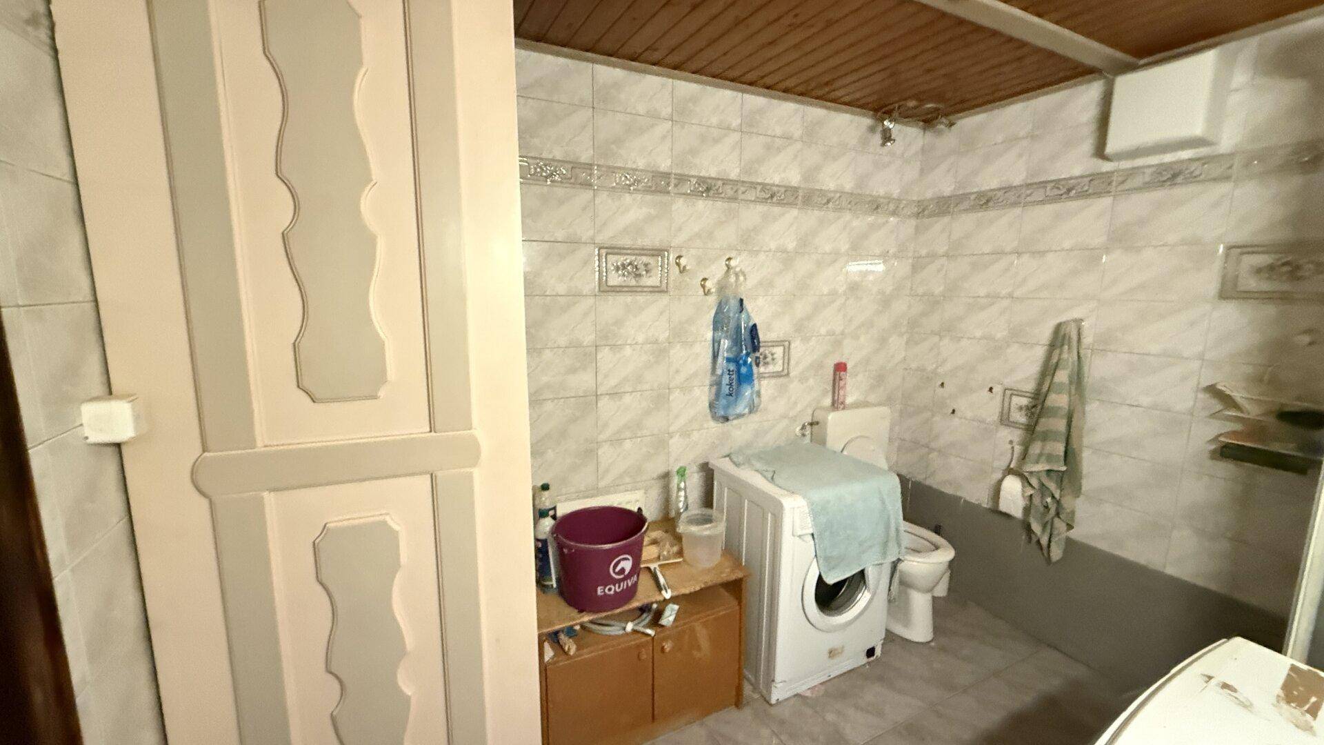 Einfaches Badezimmer mit Waschmaschine, WC und gefliesten Wänden unter einer Holzdecke.