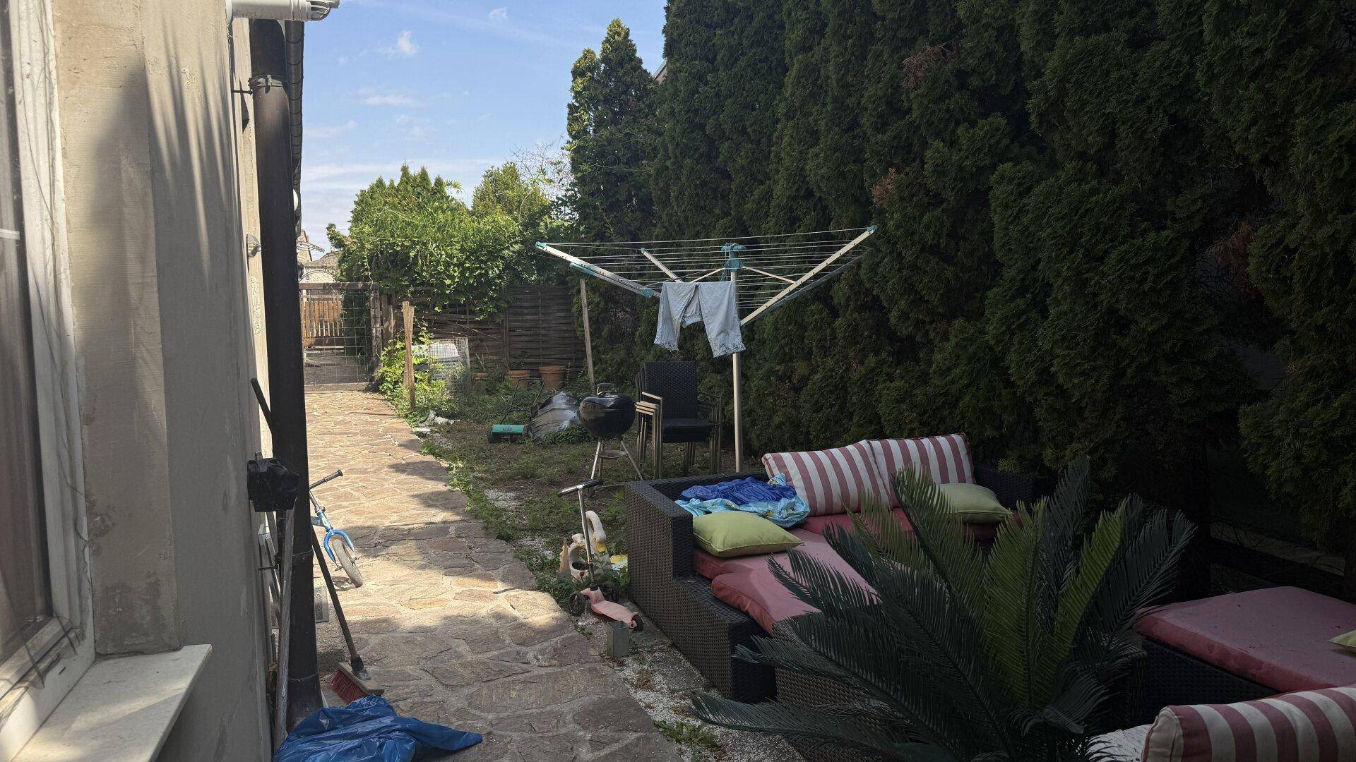 Gartenbereich mit Wäscheständer und Lounge-Möbeln, umgeben von hohen Hecken und Bäumen.