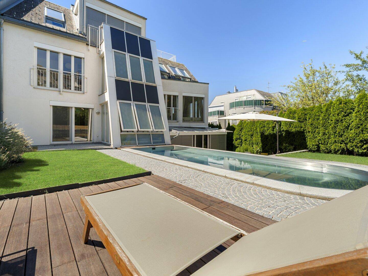 Entspannungsbereich im Garten mit Holzdeck, Liegestühlen und Pool neben der modernen Hausfassade.
