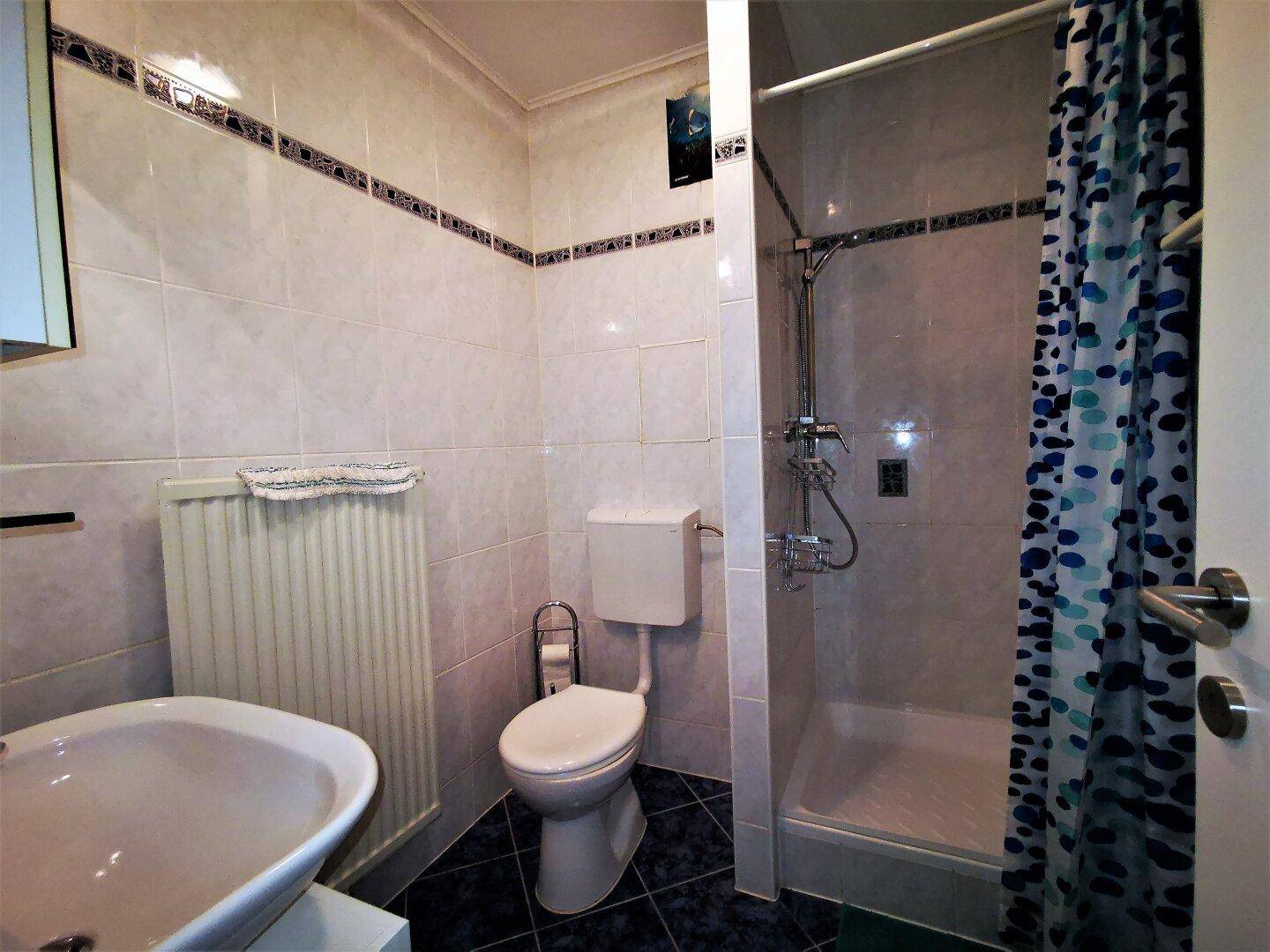 Gefliestes Badezimmer mit Dusche, WC, Waschbecken und Heizkörper, in gutem Zustand.