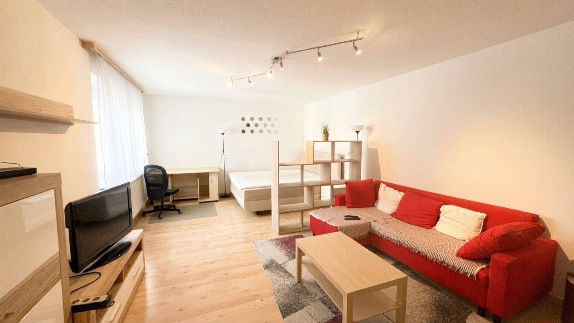 Helles Studio-Apartment mit rotem Sofa, Bett und Schreibtisch in einem offenen Wohnbereich.