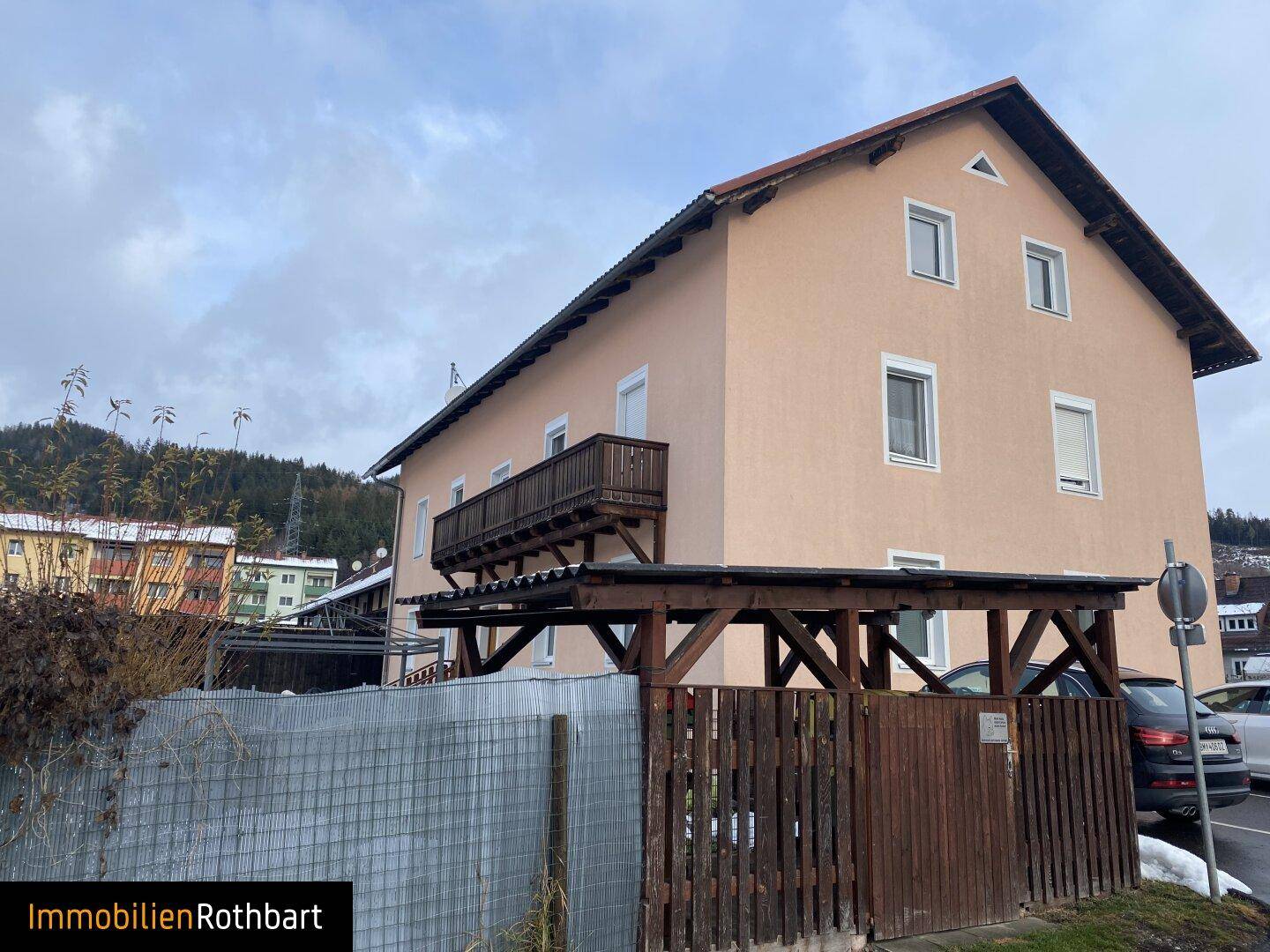 Seitenansicht des Gebäudes mit einem hölzernen Balkon und einem Carport im Vordergrund.