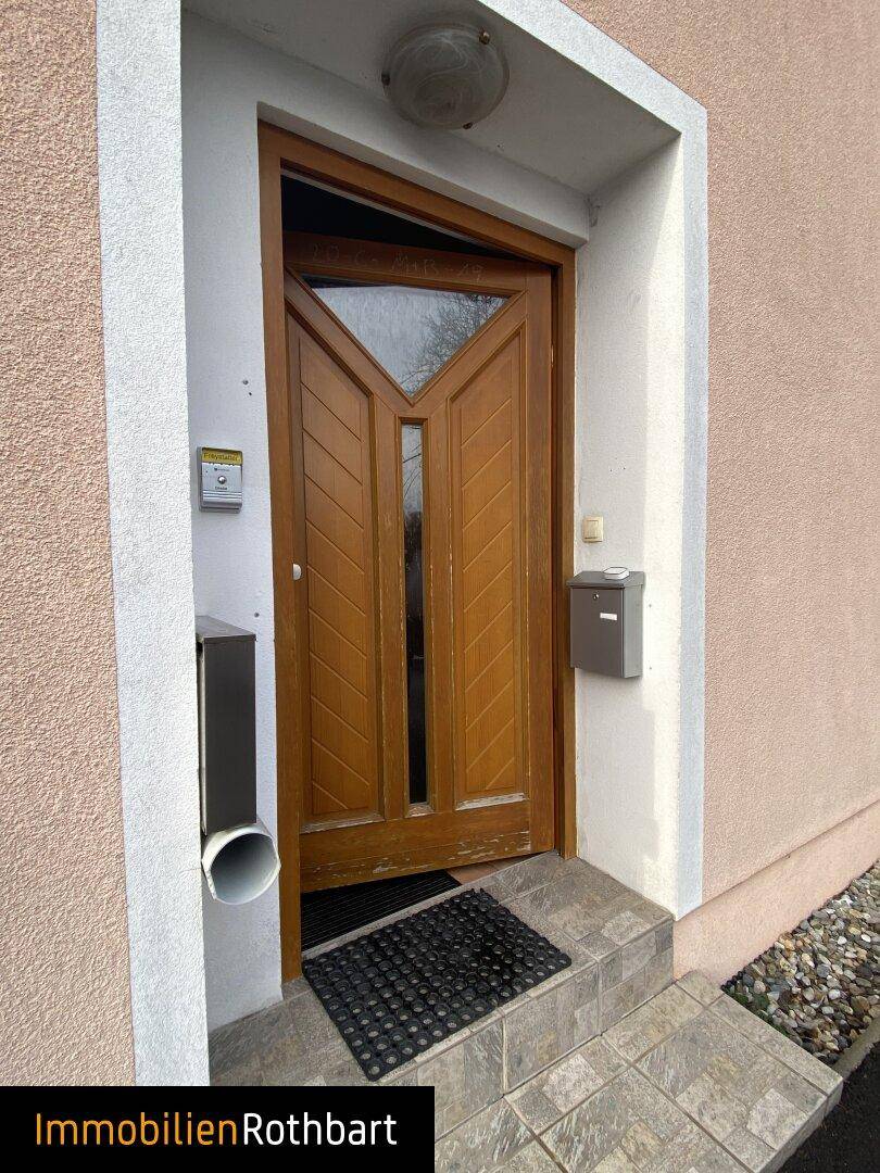 Detailansicht des Hauseingangs mit einer Holztür und einer kleinen Treppe, umgeben von Putzfassade.