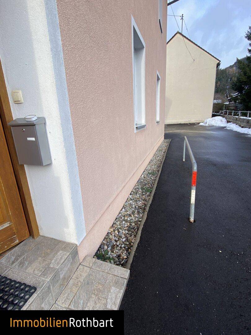 Seitenansicht des Gebäudes mit einem schmalen Kiesstreifen entlang der Wand und einer Zufahrt.