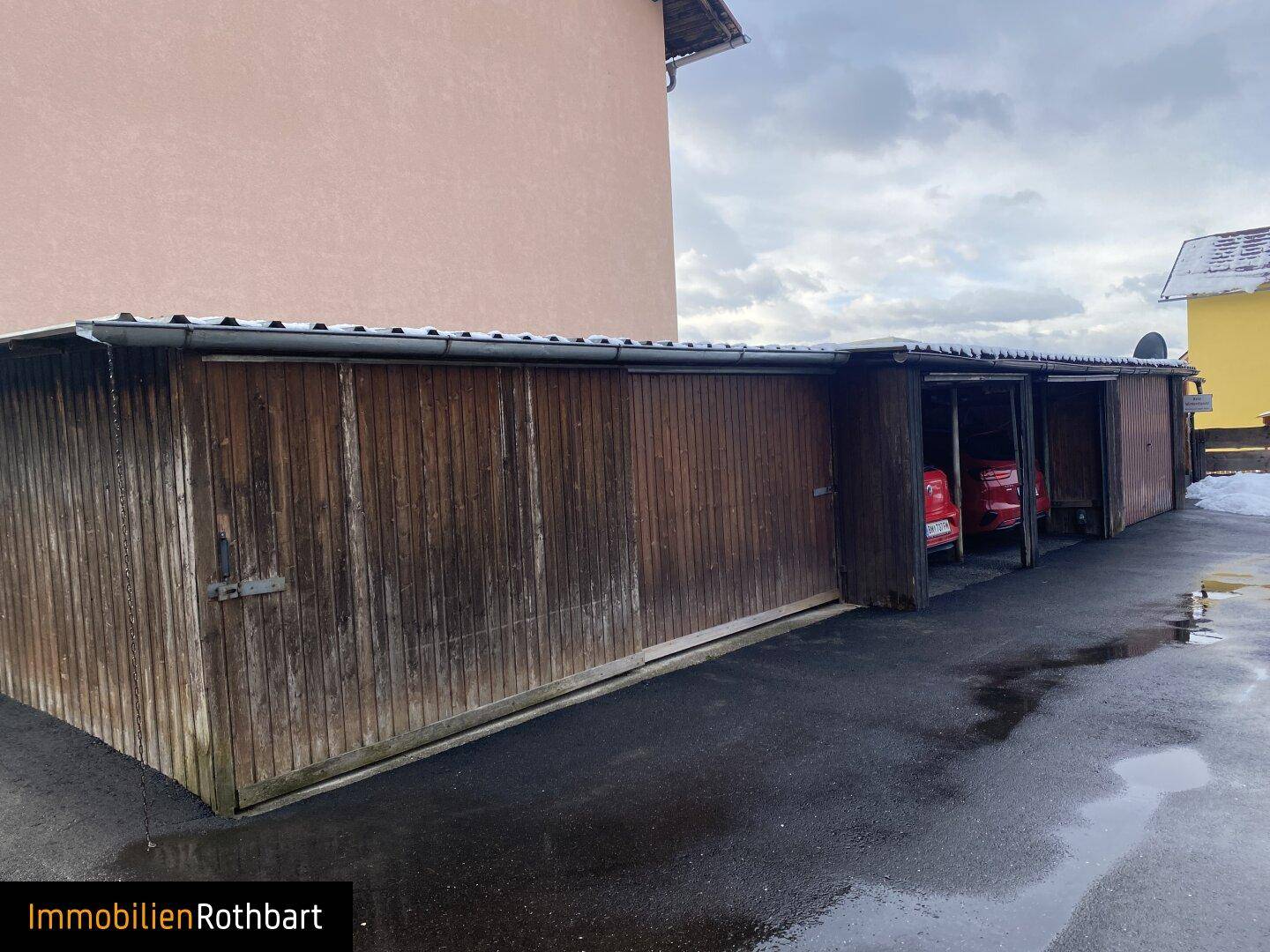 Reihe von Holzgaragen mit Schiebetoren und einem gepflasterten Vorplatz, teilweise mit Schnee bedeckt.