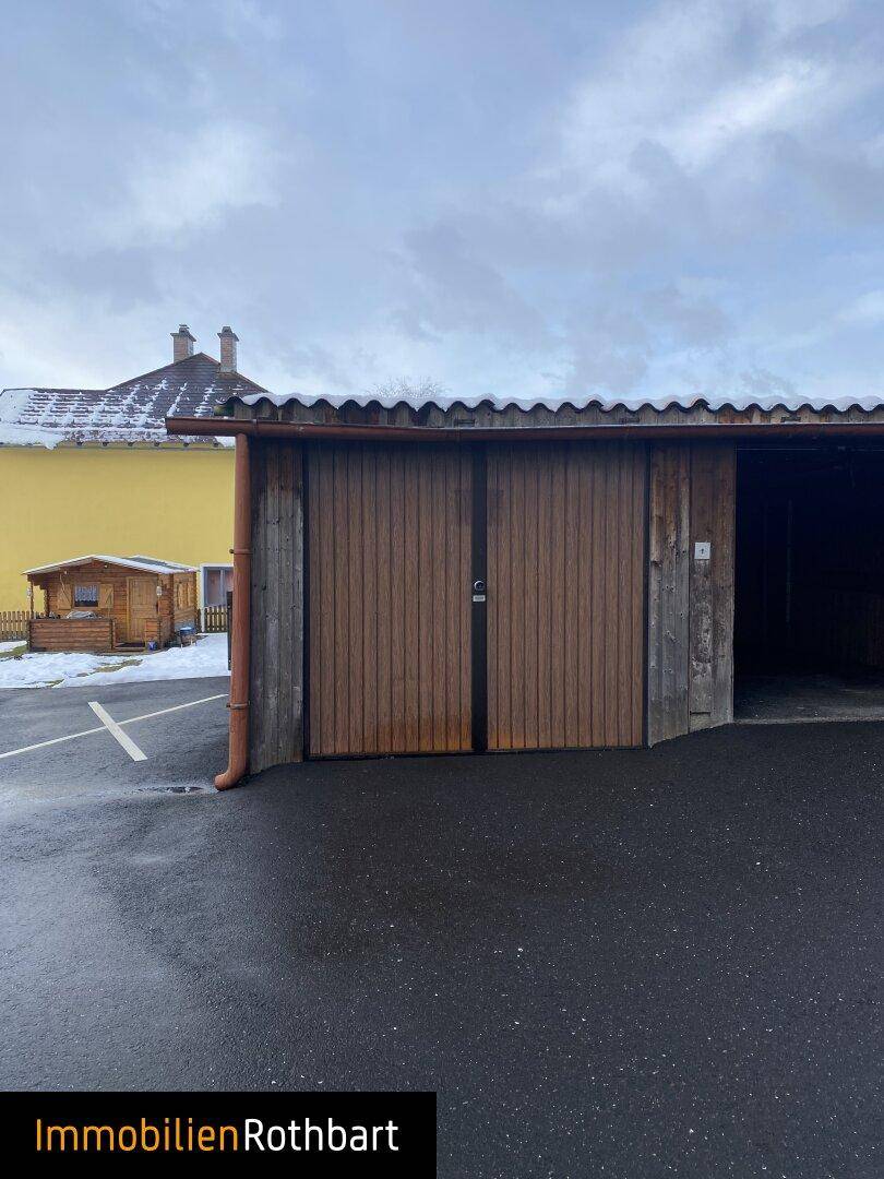 Zwei Holzgaragen mit braunen Toren und einem asphaltierten Vorplatz, im Hintergrund ein weiteres Gebäude.