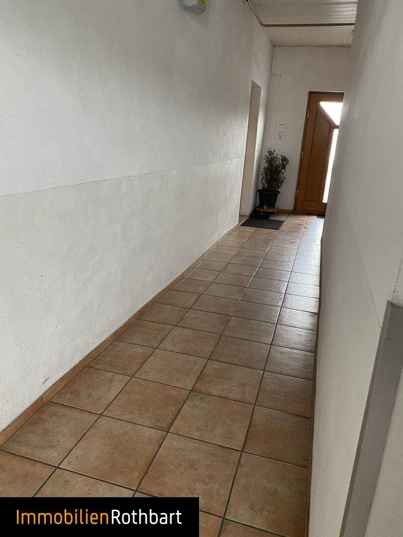 Langer Flur mit hellbraunen Fliesen und weißen Wänden, der zu einer Holztür am Ende führt.