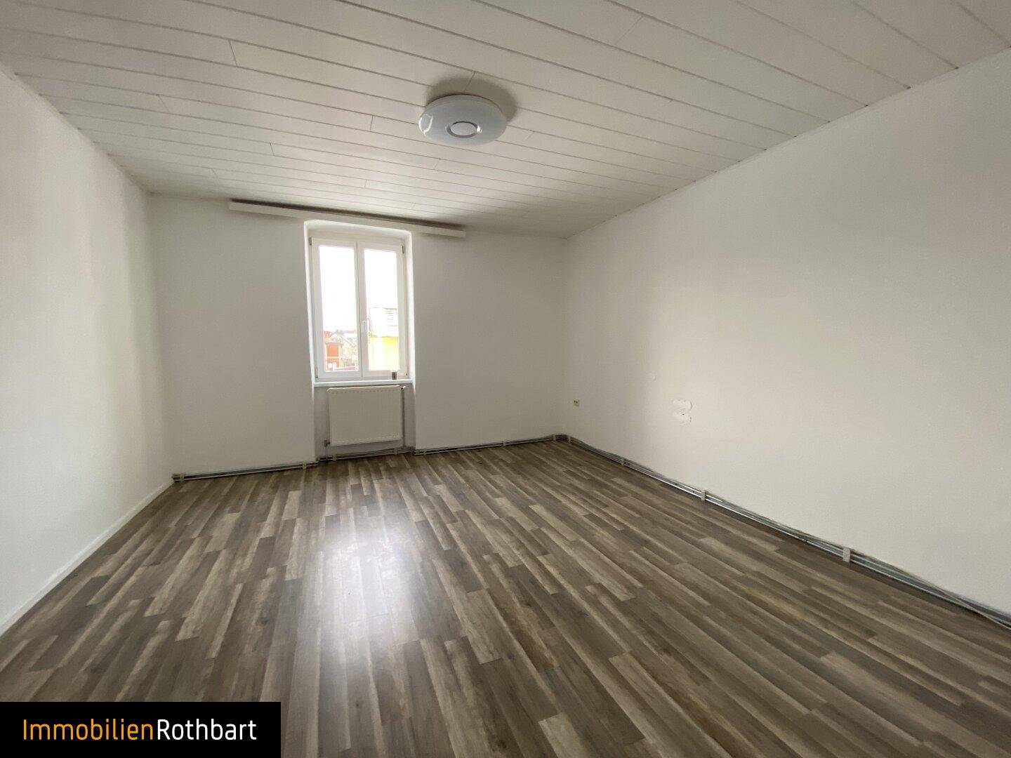 Helles Zimmer mit Laminatboden, weißen Wänden und einer Fensterfront, die viel Tageslicht hereinlässt.