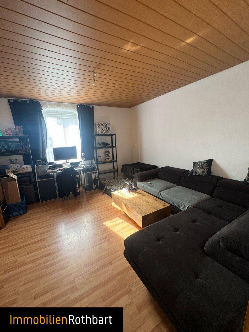 Wohnzimmer mit großem Sofa, Holztisch und Laminatboden, durch ein Fenster fällt Tageslicht herein.