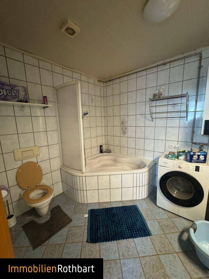 Badezimmer mit Eckbadewanne, Toilette und Waschmaschine, gefliest in hellen Farben.