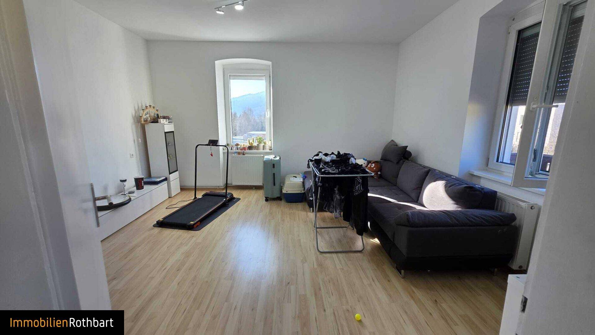 Geräumiges Wohnzimmer mit hellem Holzboden, Sofa und Blick auf die Berge durch das Fenster.