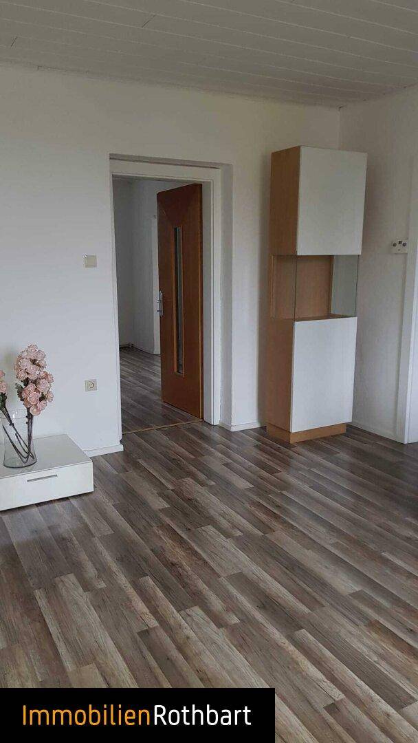 Helles Zimmer mit Laminatboden und weißen Wänden, ein offener Durchgang führt zu einem weiteren Raum.