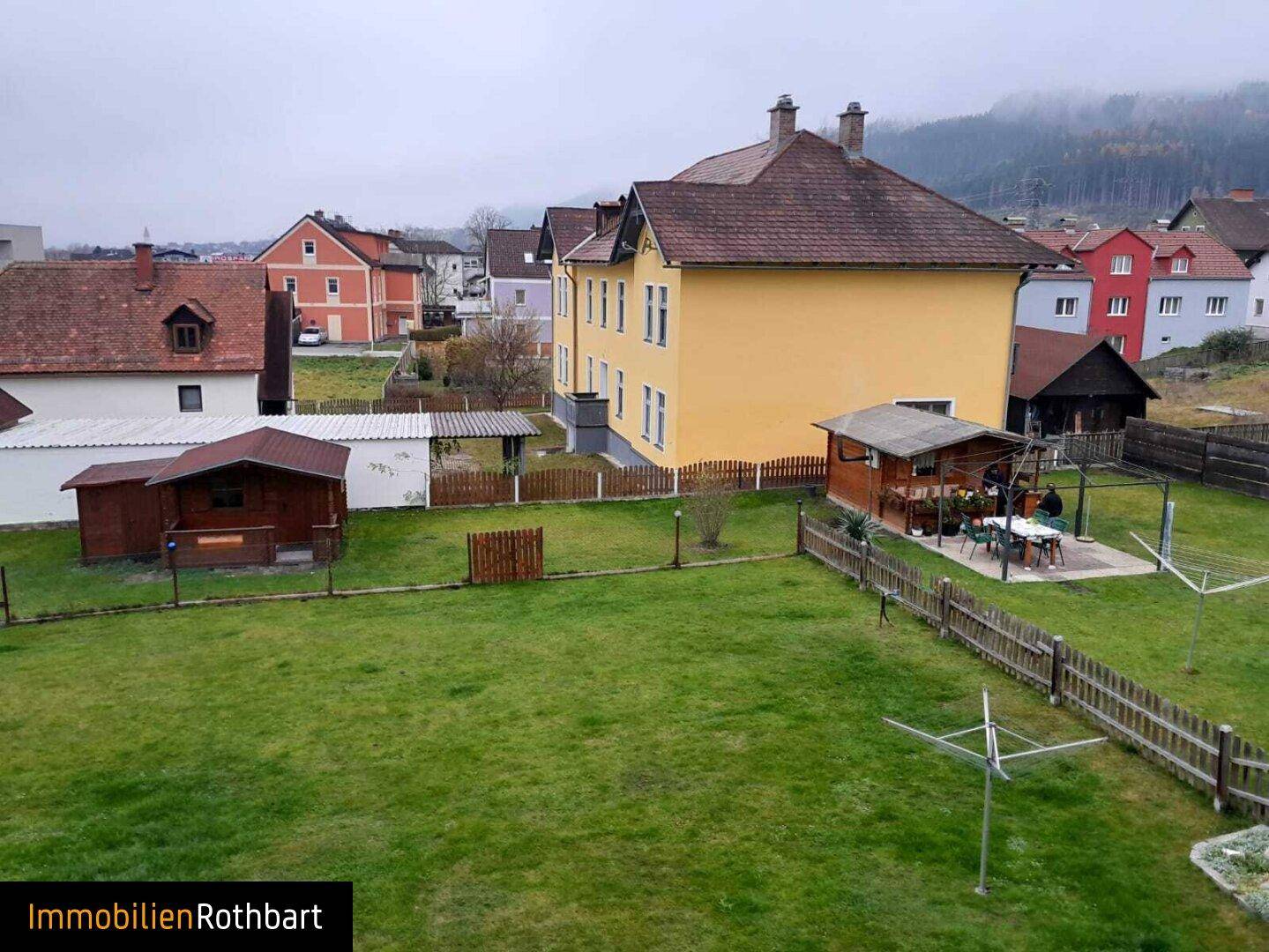 Weitläufiger Gartenbereich mit Rasenfläche, kleinen Gartenhäusern und Blick auf umliegende Gebäude und Berge.