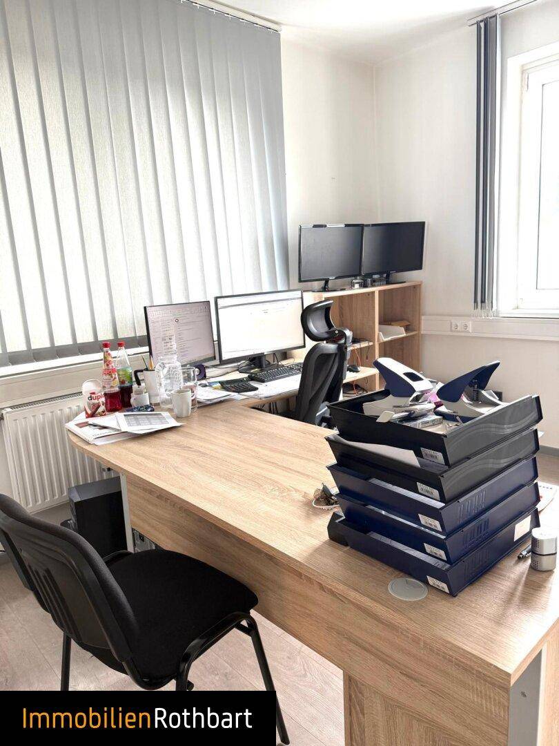 Büroarbeitsplatz mit Holzschreibtisch, zwei Monitoren und vertikalen Jalousien am Fenster.
