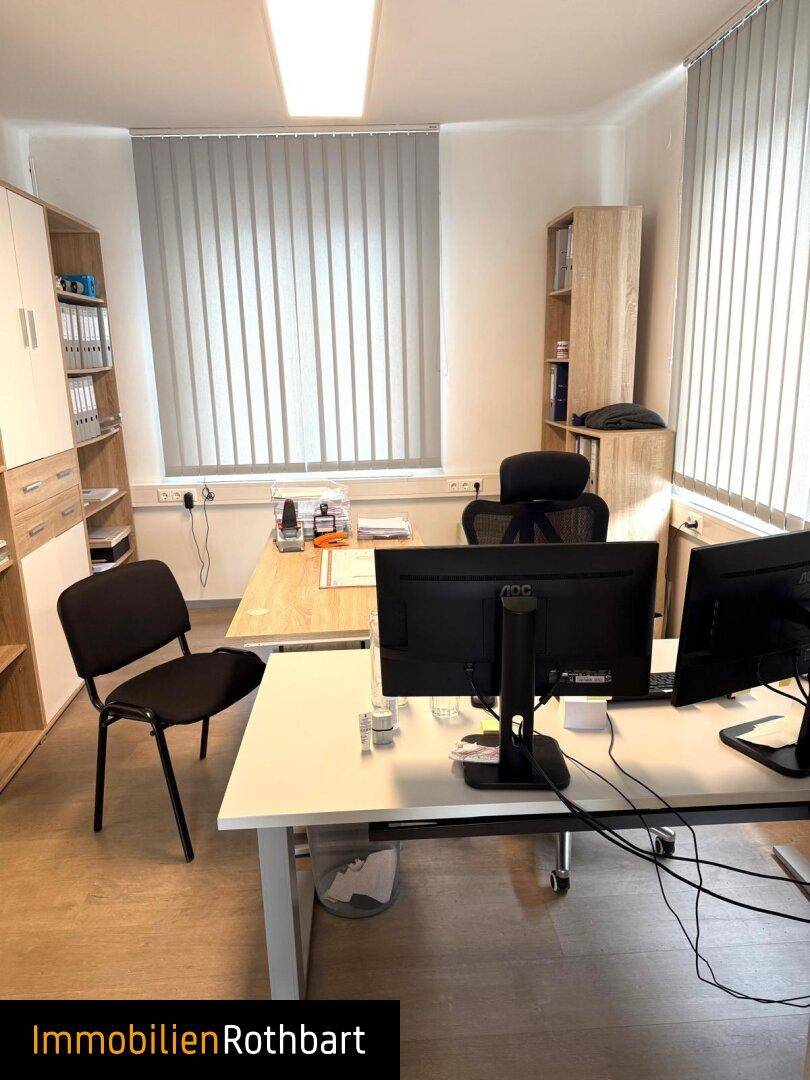 Geräumiges Büro mit mehreren Arbeitsplätzen, Regalen und vertikalen Fensterjalousien.