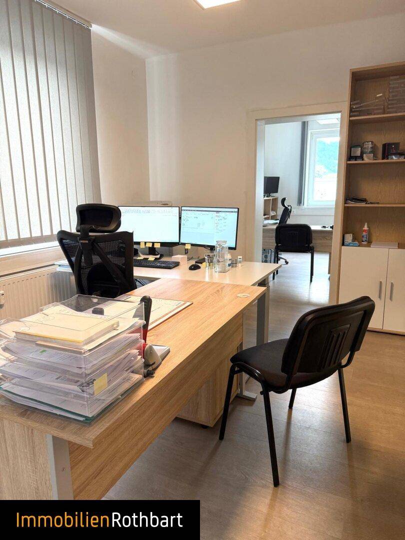 Büro mit Holzschreibtisch und Blick in einen weiteren Arbeitsbereich durch die Türöffnung.