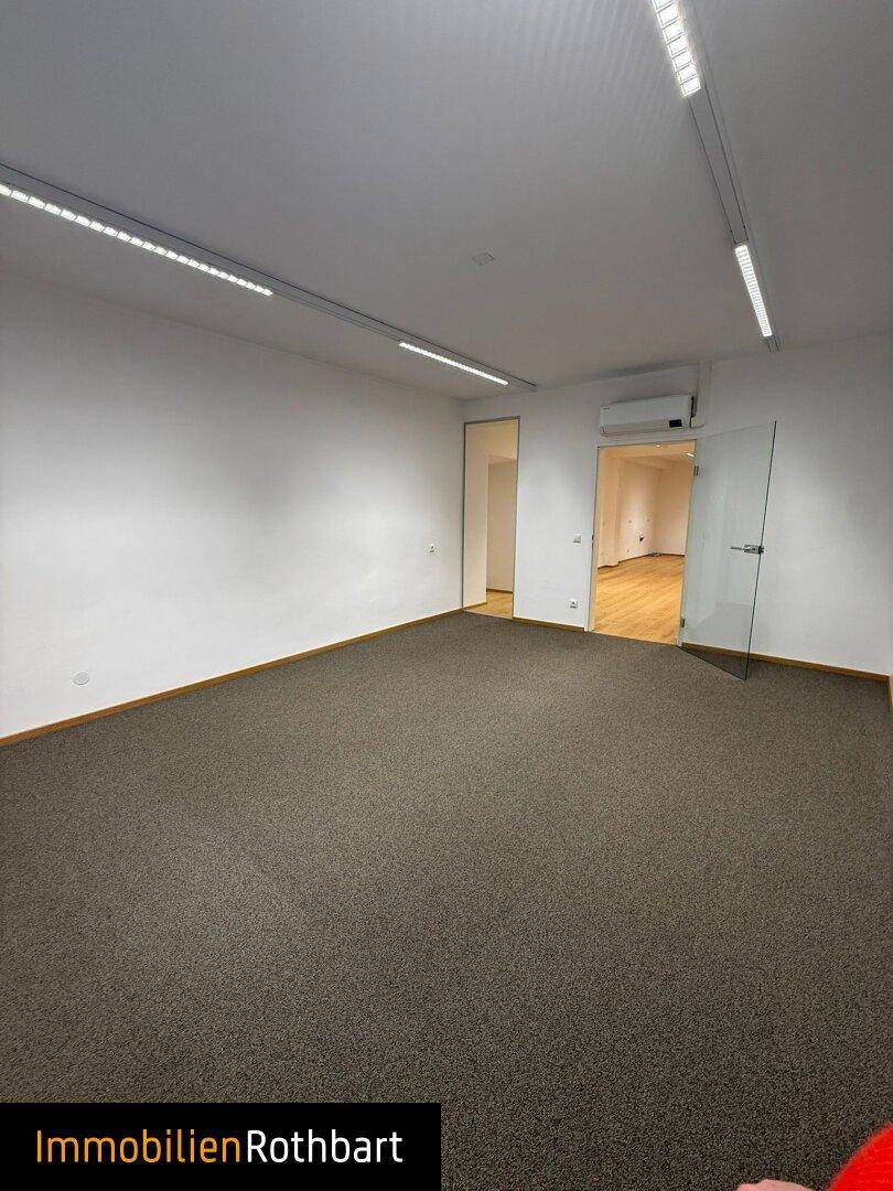 Geräumiger Büroraum mit Teppichboden, weißen Wänden und Deckenbeleuchtung.