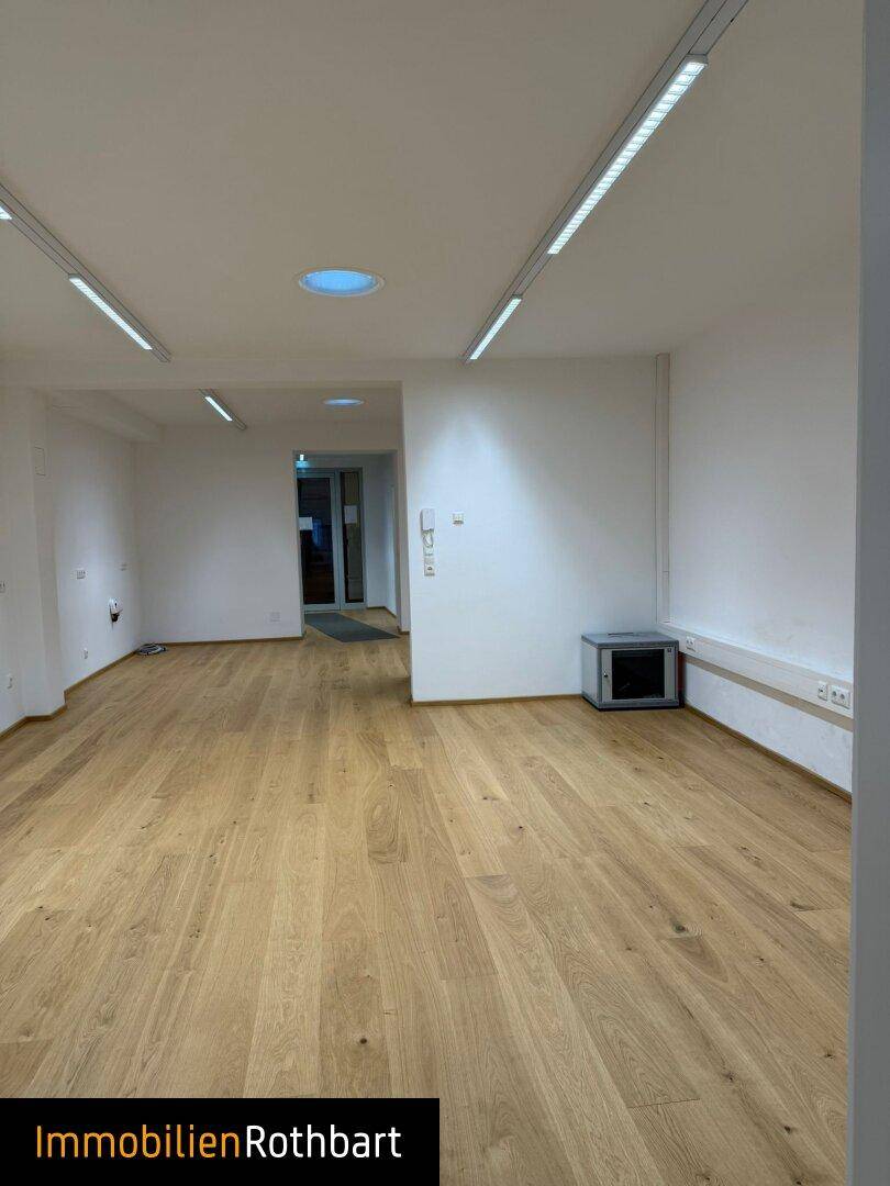 Leerer Büroraum mit Holzboden, weißen Wänden und Deckenleuchten, ideal für flexible Nutzung.