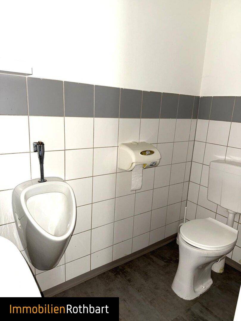 Einfache Gästetoilette mit Urinal und WC, gefliest in Weiß und Grau, mit Toilettenpapierspender.