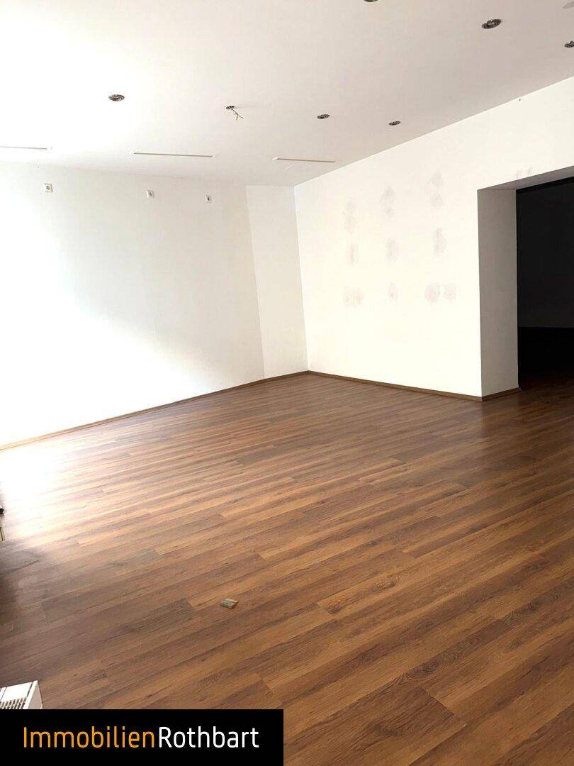 Weiter Blick in den leeren Geschäftsraum mit Laminatboden und einem Durchgang zu einem weiteren Bereich.