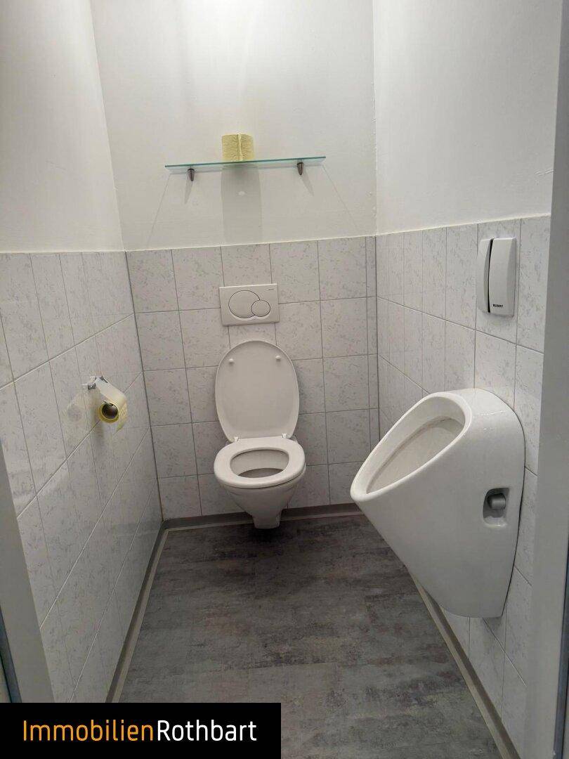 Kleine Gästetoilette mit WC und Urinal, halbhoch gefliesten Wänden und modernem grauen Boden.