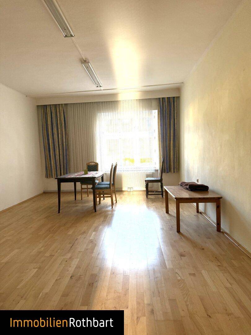 Heller Wohnraum mit Holzboden, Vorhängen und zwei kleinen Tischen am Fenster.