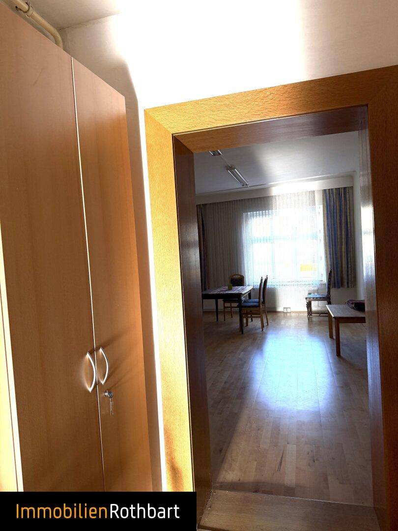 Flur mit Holzschrank und Blick in einen hellen Raum mit Fenstern.