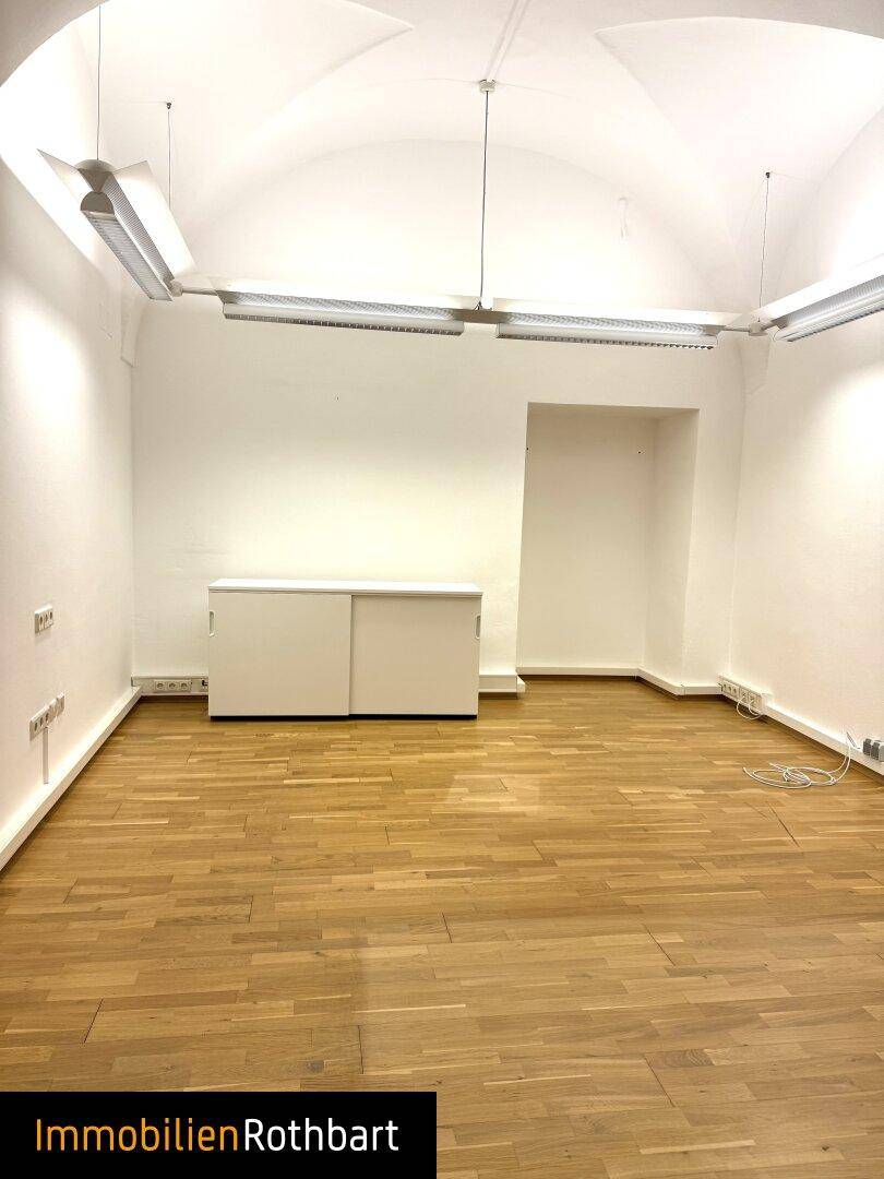 Leerer Büroraum mit Holzboden, gewölbter Decke und einem weißen Sideboard an der Wand.