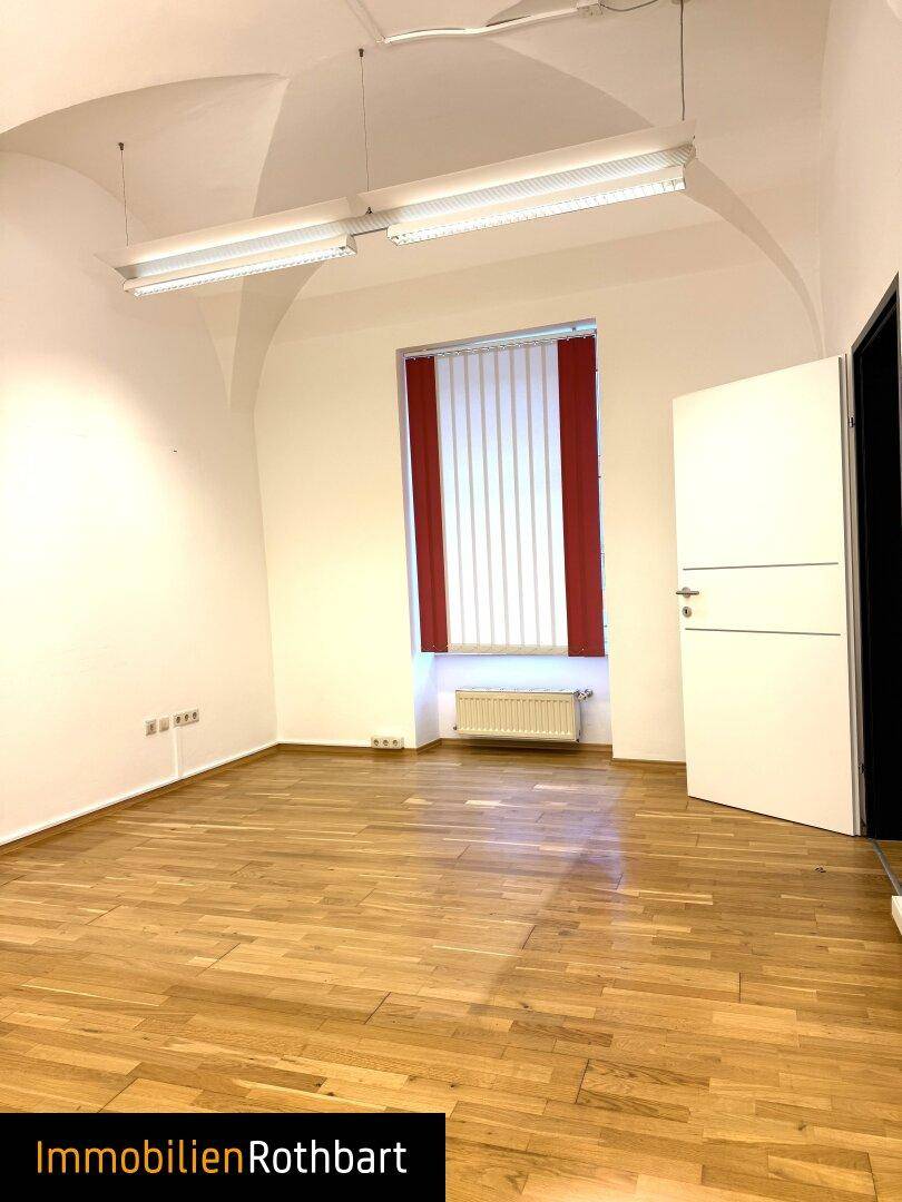 Heller Büroraum mit Holzboden, gewölbter Decke und einem Fenster mit roten Jalousien.