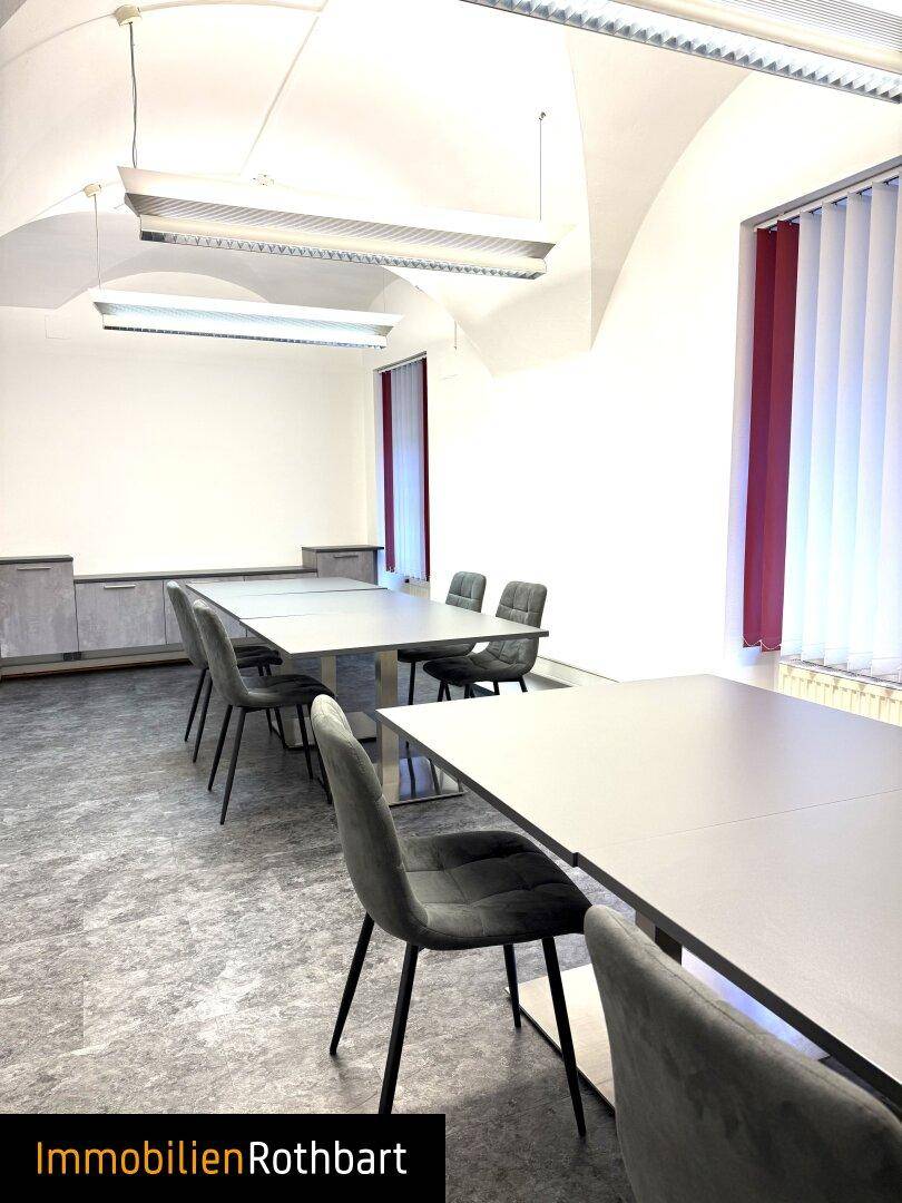 Moderner Konferenzraum mit gewölbter Decke, langen Tischen und vertikalen Jalousien an den Fenstern.