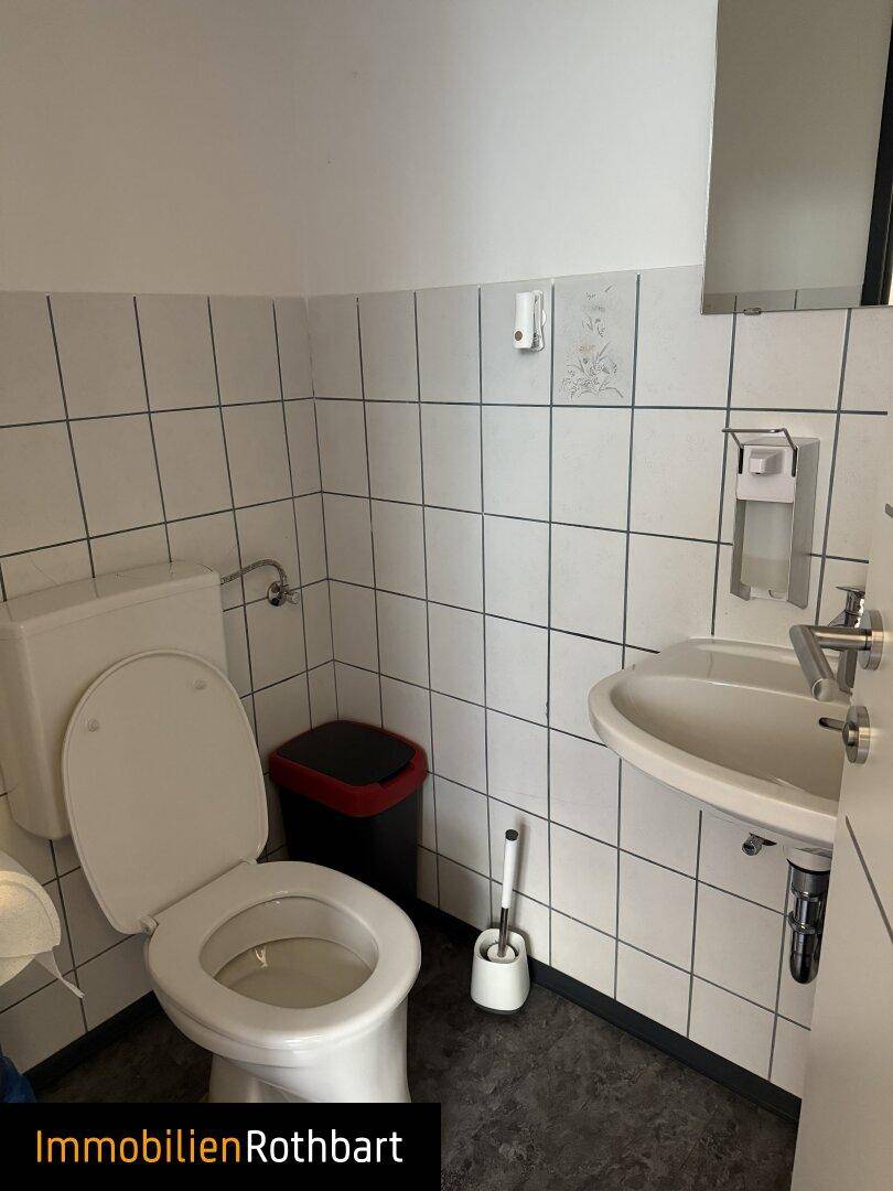 Einfache Toilette mit weißen Fliesen, einem Waschbecken und einem kleinen Mülleimer im Eckbereich.