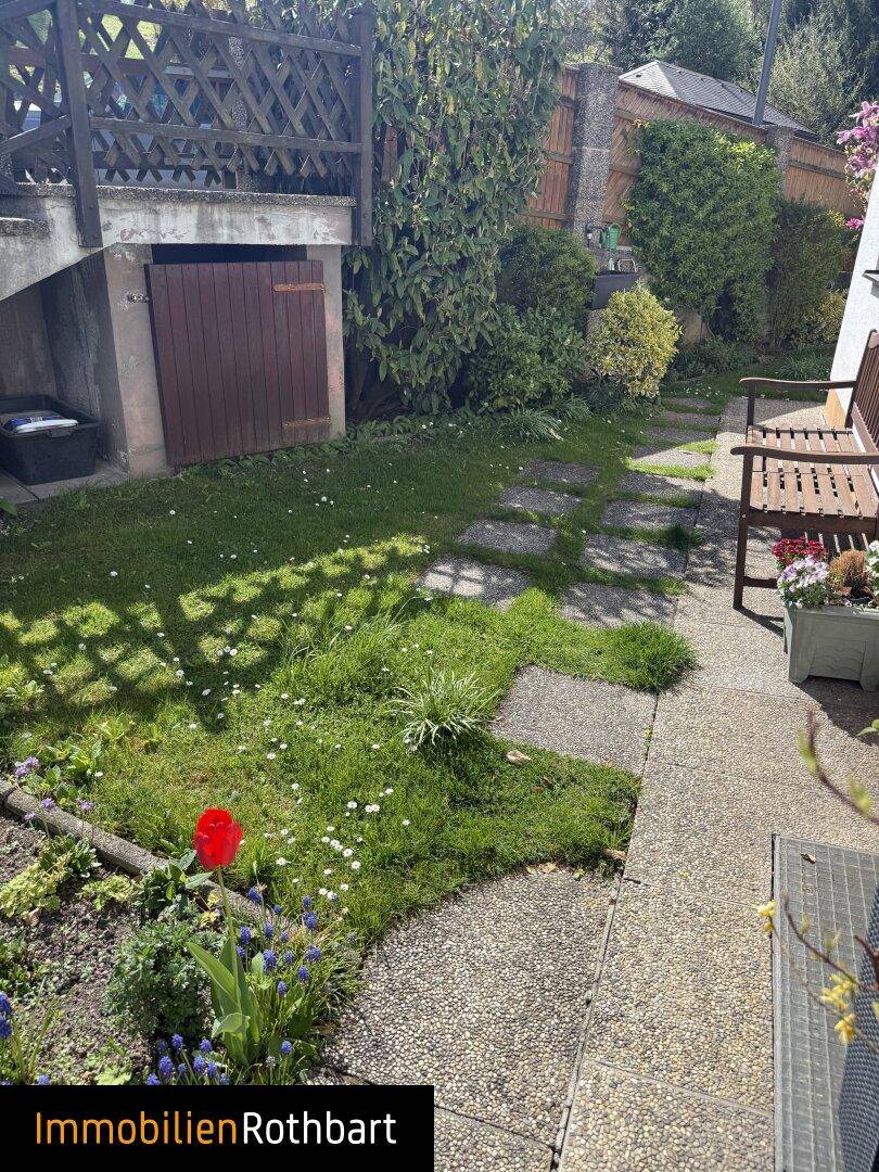 Gepflegter Gartenweg mit Blumen und einer kleinen Holztür zu einem Lagerbereich.