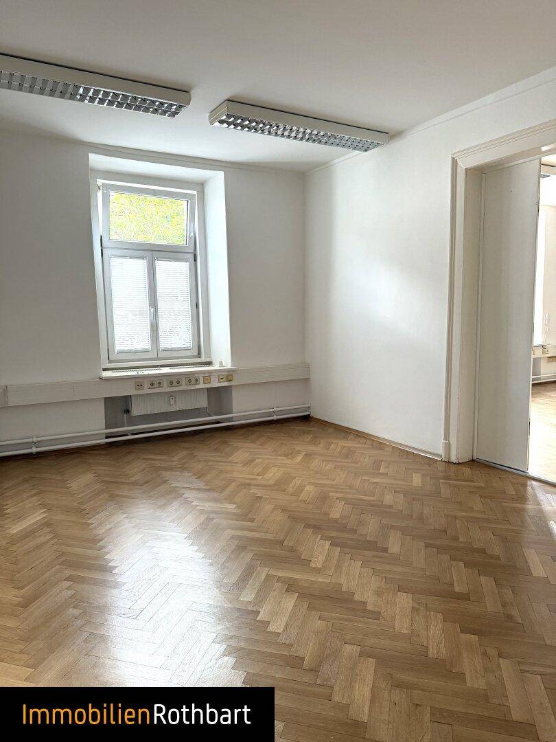 Geräumiger Büroraum mit elegantem Fischgrätparkett, weißen Wänden und einem großen Fenster.