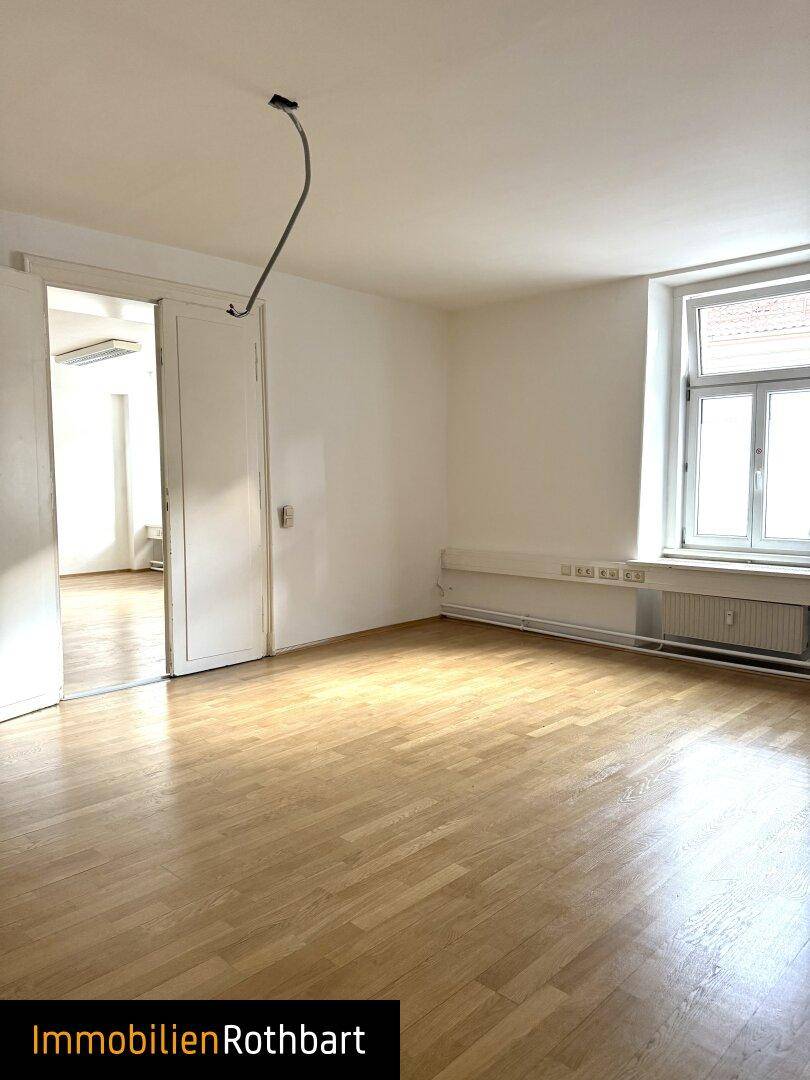 Geräumiger Raum mit Holzparkett, weißen Wänden, Fenster und Zugang zu einem Nebenraum.