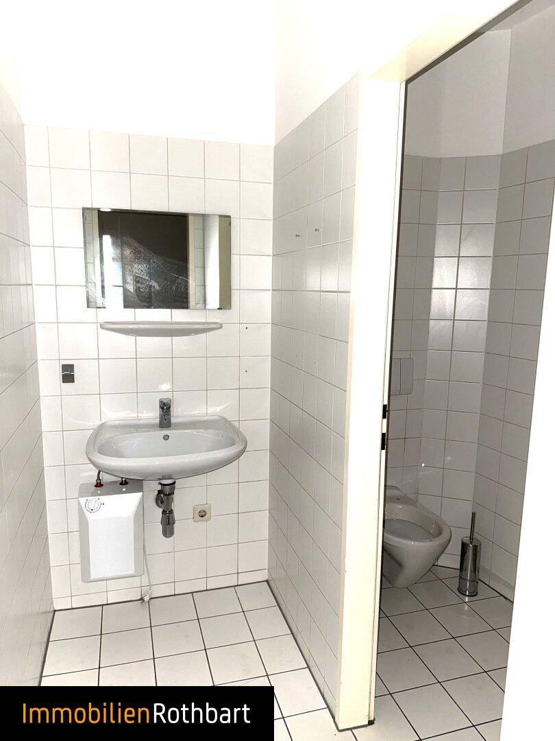 Badezimmer mit weißen Fliesen, Waschbecken, Spiegel und separatem WC-Bereich.