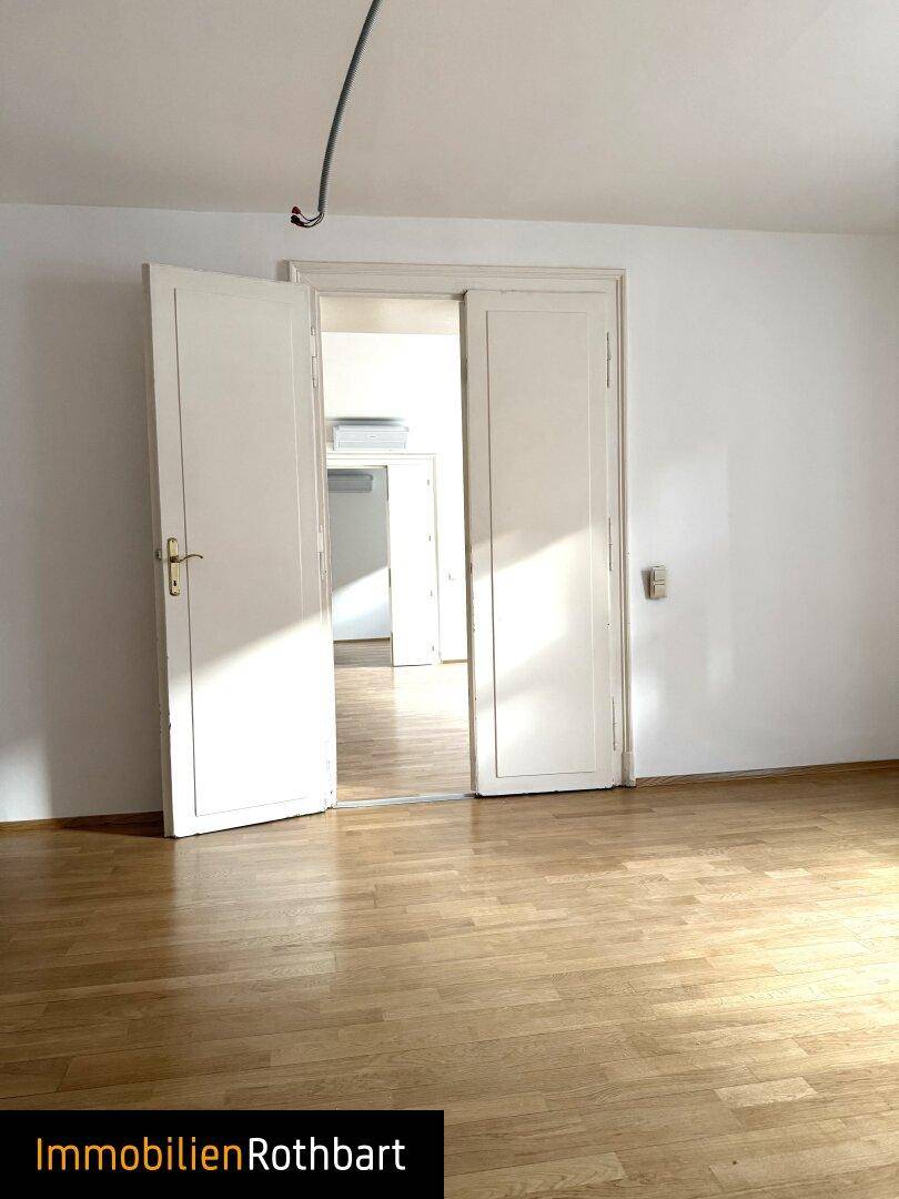 Heller Raum mit Holzparkett, weißen Wänden und einer weit geöffneten Doppeltür.