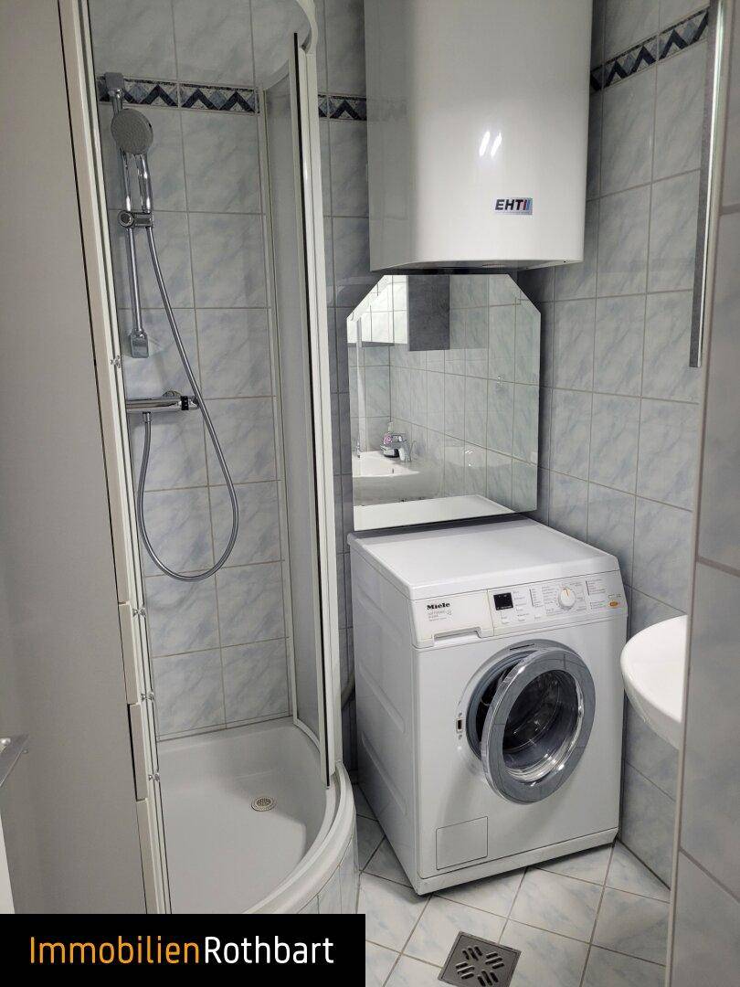 Funktionales Badezimmer mit Duschkabine, Waschmaschine und kleinem Waschbecken. Helle Fliesen an Wänden und Boden.