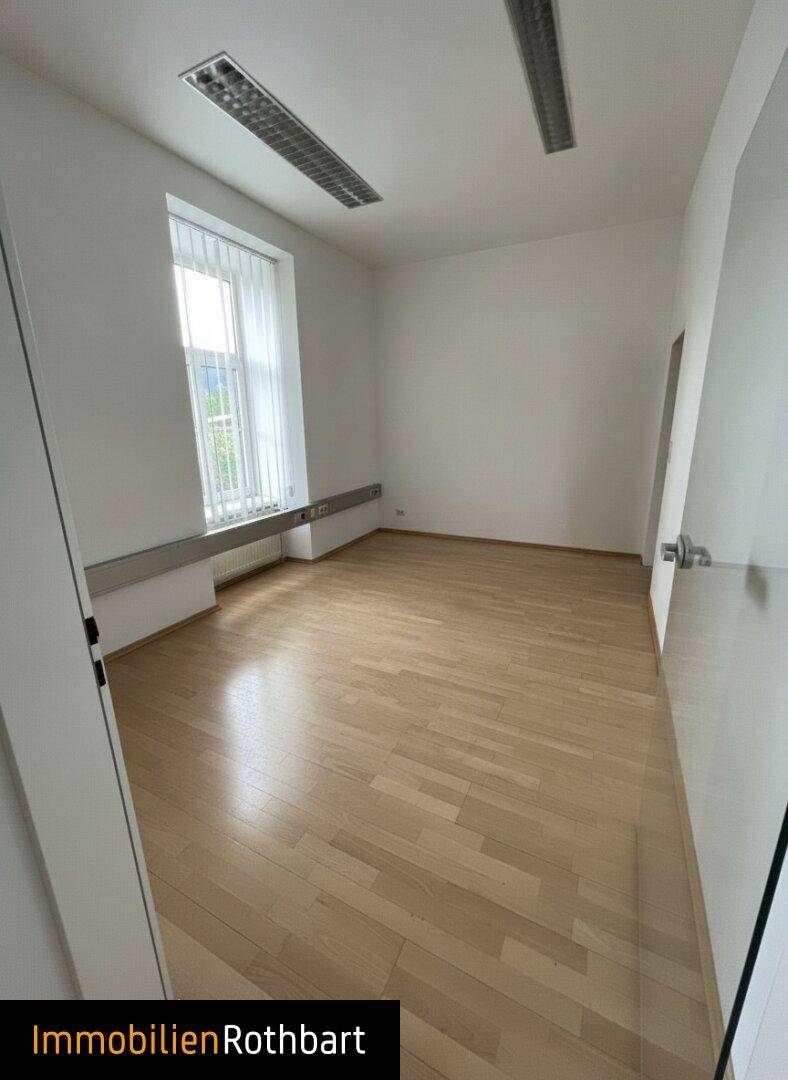 Heller Büroraum mit Holzfußboden, weißen Wänden und einem großen Fenster, das viel Tageslicht hereinlässt.