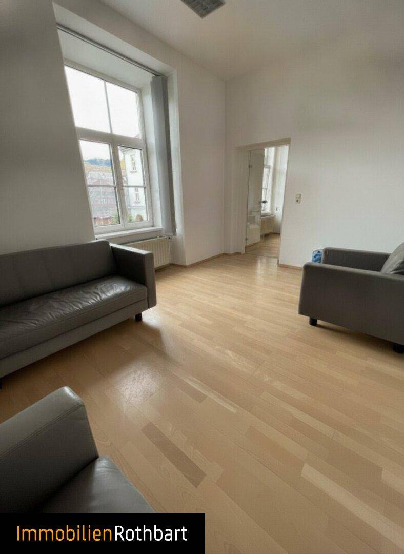 Gemütlicher Raum mit hellem Holzfußboden, weißen Wänden und zwei grauen Sofas, ideal als Wartebereich oder Lounge.