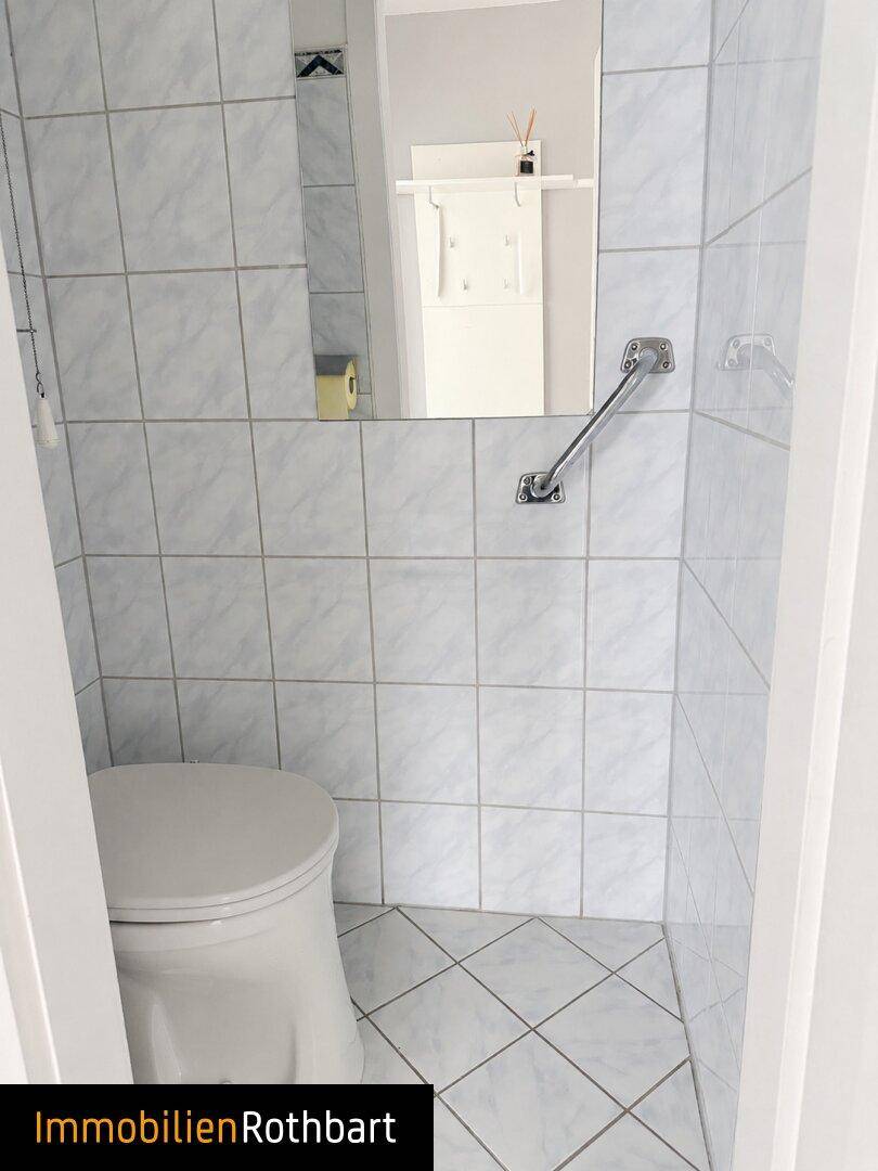 Separates WC mit Toilette, Haltegriff und hellen Fliesen an den Wänden.