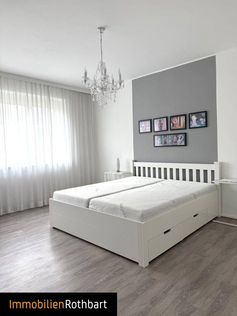 Helles Schlafzimmer mit weißem Bett, grauer Akzentwand und Kronleuchter. Große Fenster sorgen für viel Licht.