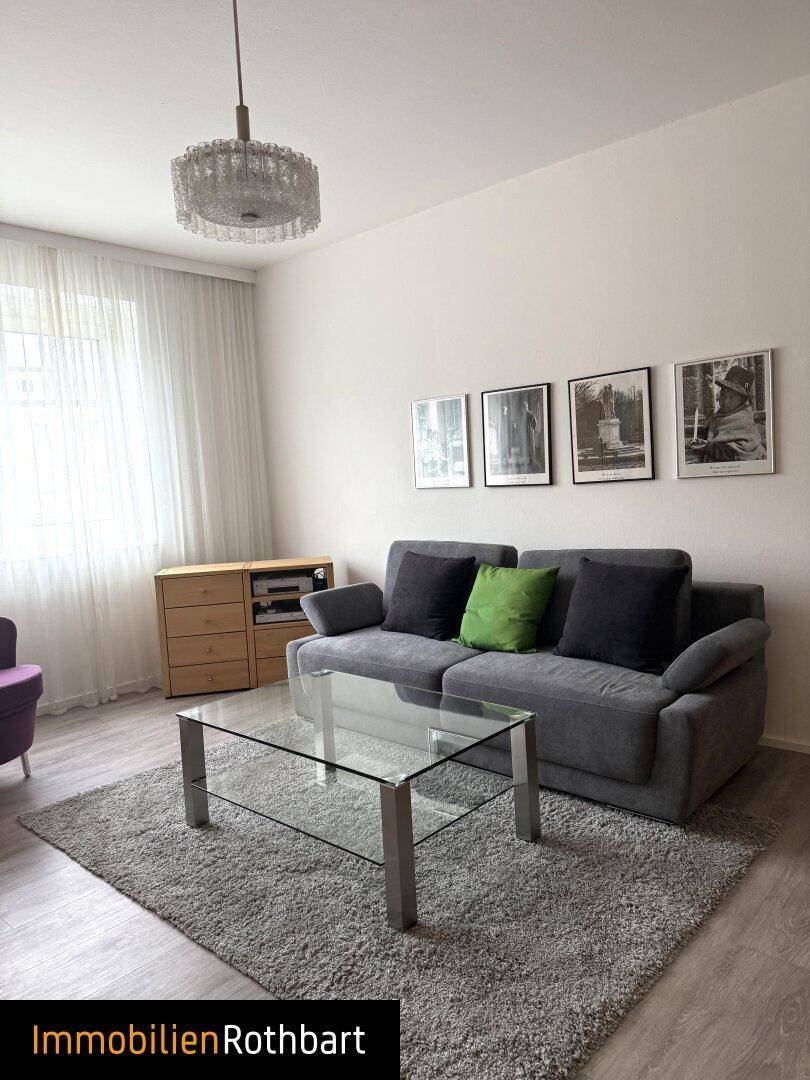Gemütliches Wohnzimmer mit grauem Sofa, Glastisch und Laminatboden. Helle Wände und Fenster.