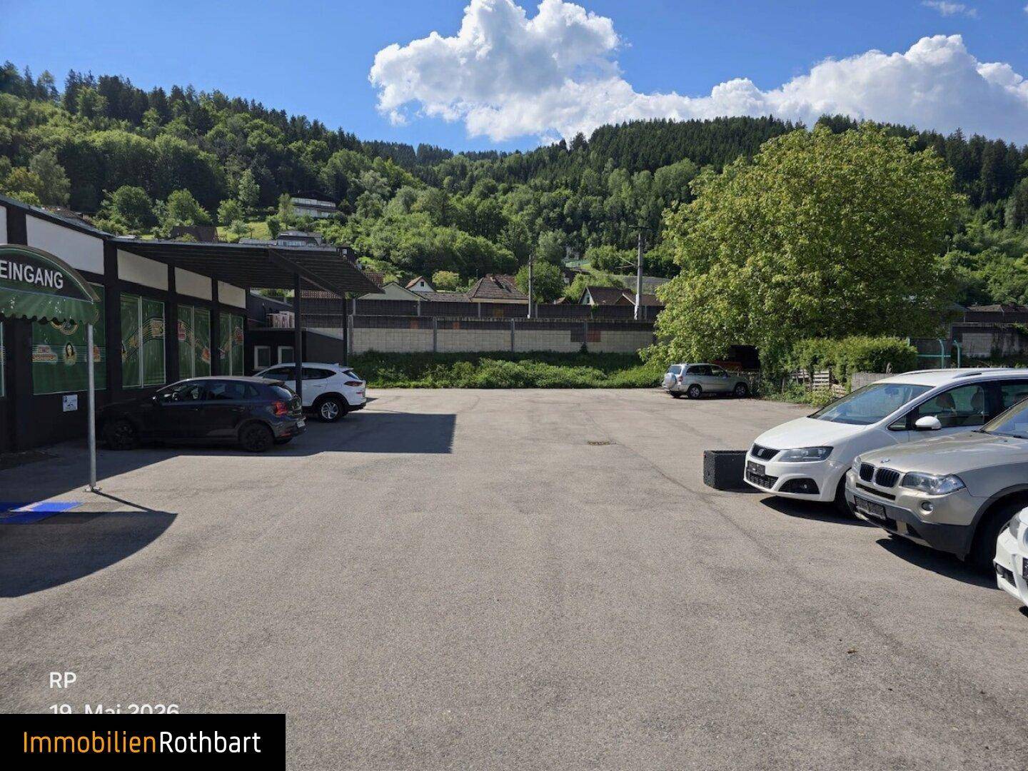 Großer Parkplatz vor dem Gebäude mit Blick auf die umliegende grüne Natur und Bäume im Sonnenschein.