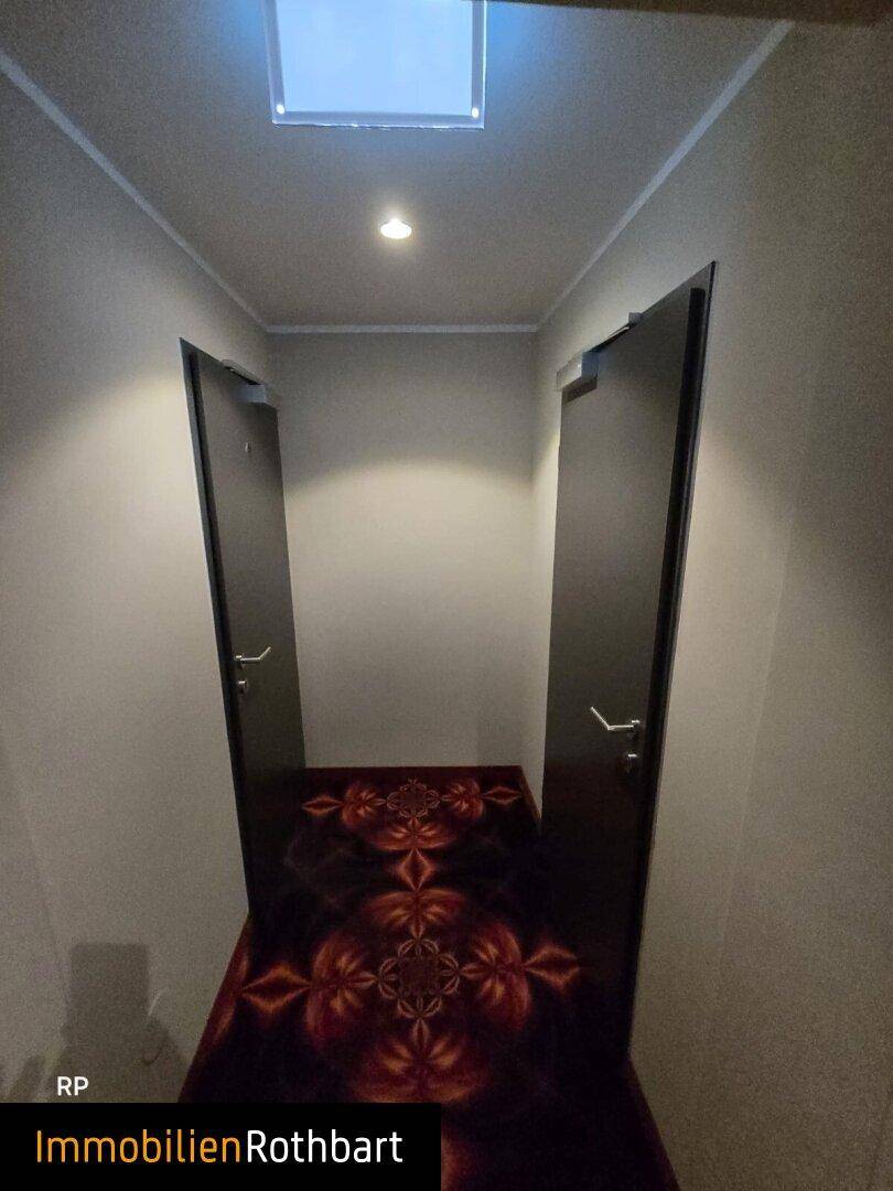Schmaler Flur mit gemustertem Teppichboden und zwei Türen, beleuchtet von Deckenleuchten.