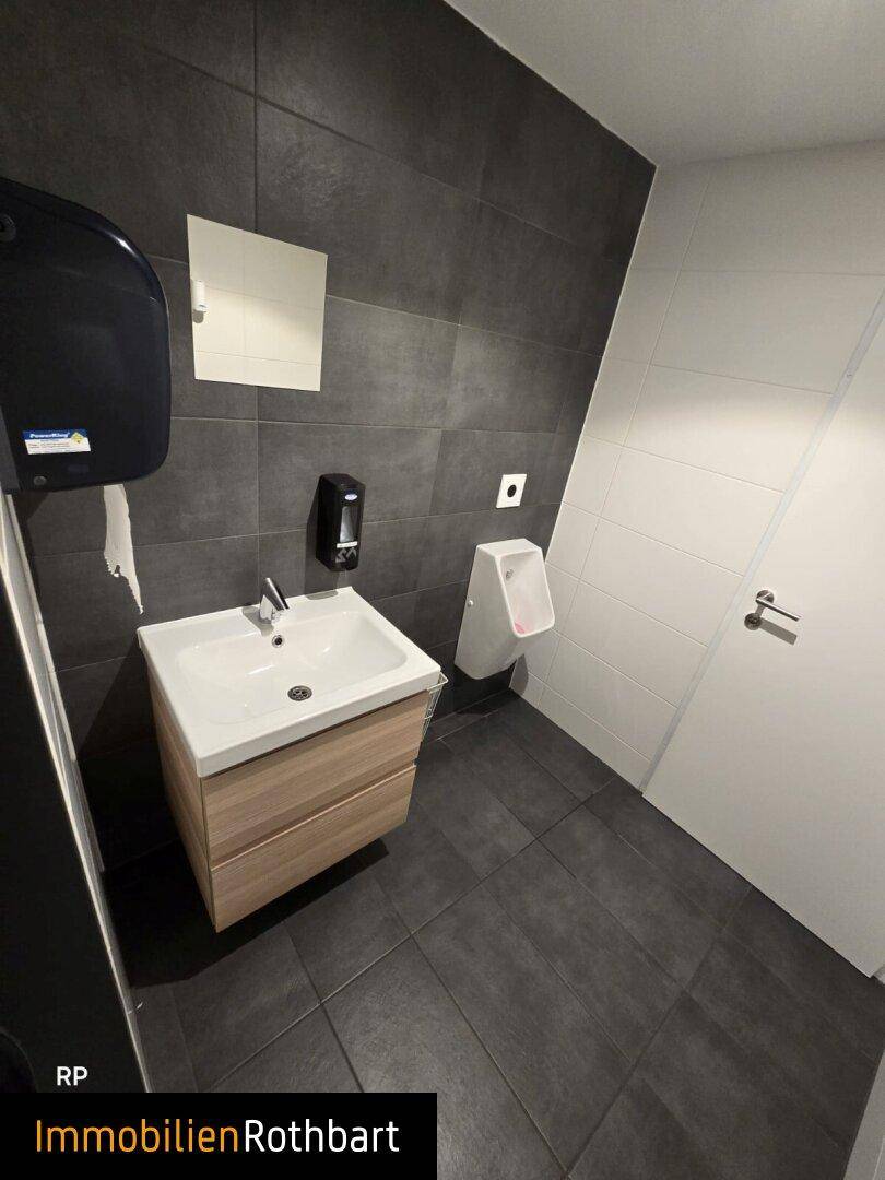 Moderner Toilettenraum mit Waschbecken, Urinal und dunklen Fliesen an Wänden und Boden.