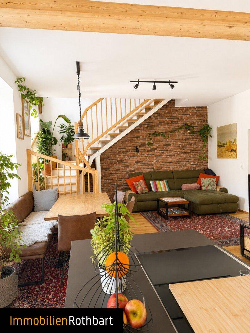 Offener Wohn- und Essbereich mit freiliegenden Holzbalken, Ziegelwand und einer Holztreppe.