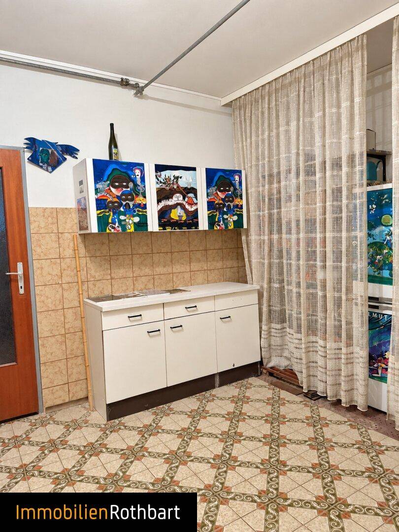 Einfache Küche mit Fliesenboden, Arbeitsplatte und Vorhängen am Fenster.