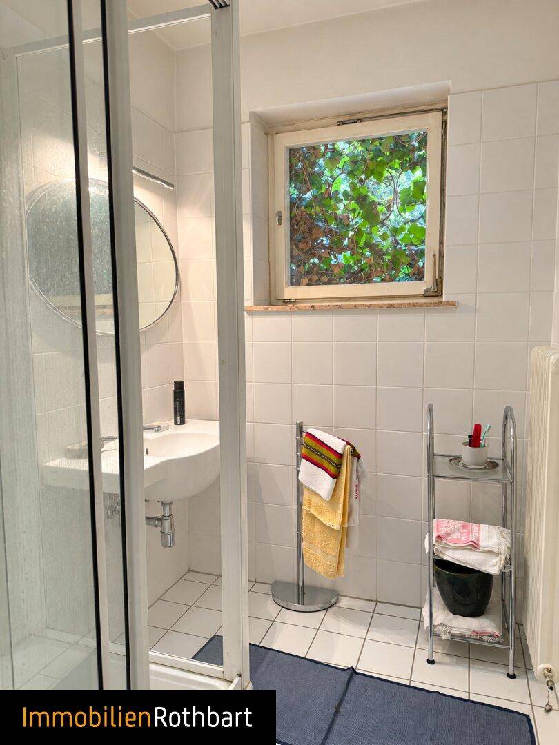 Helles Badezimmer mit Dusche, Waschbecken und Fenster, weiß gefliest.