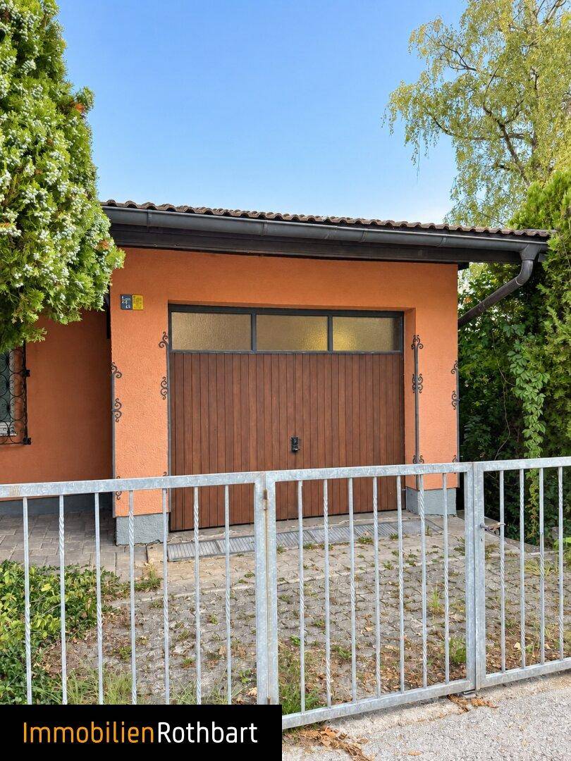 Einzelgarage mit Holztor und gepflasterter Zufahrt, umgeben von Bäumen.